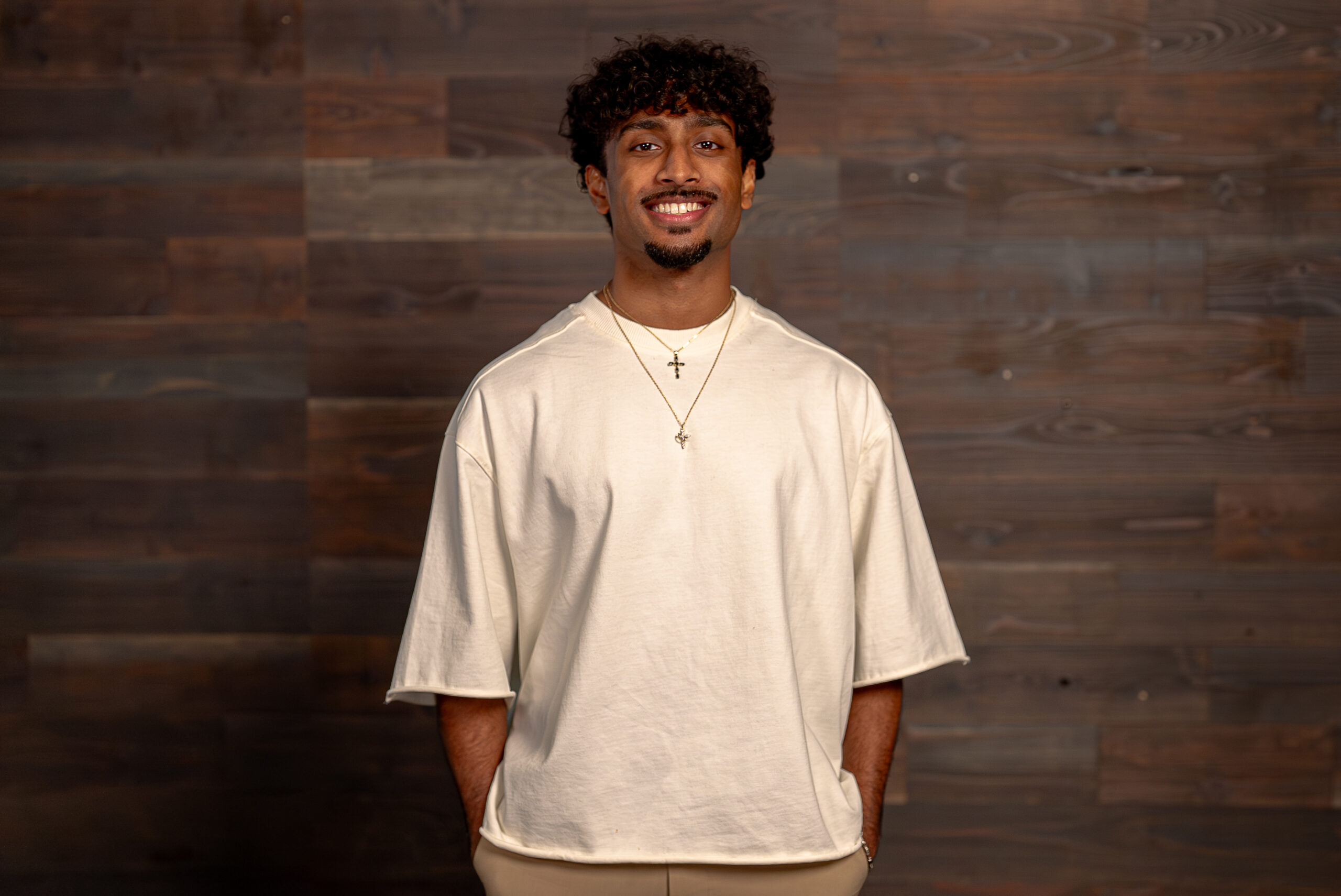 A smiling individual stands against a wooden backdrop, wearing a loose-fitting white shirt and layered gold necklaces with crosses.