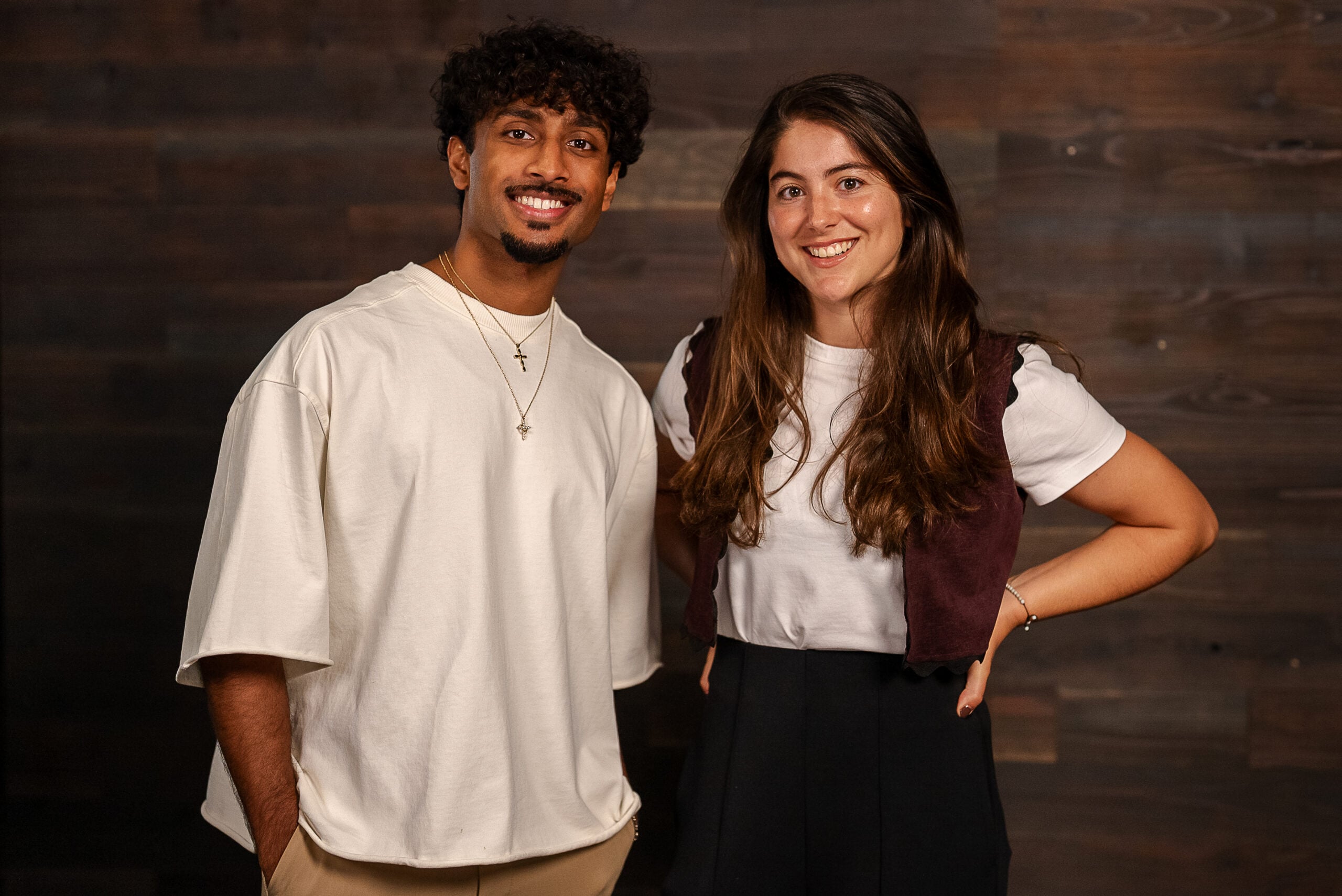 A man and a woman are smiling together in a casual setting. The man wears a loose-fitting white shirt and beige pants, accessorized with layered necklaces. The woman is dressed in a white shirt under a dark vest and black pants, with her hair down. The background features wooden panels.