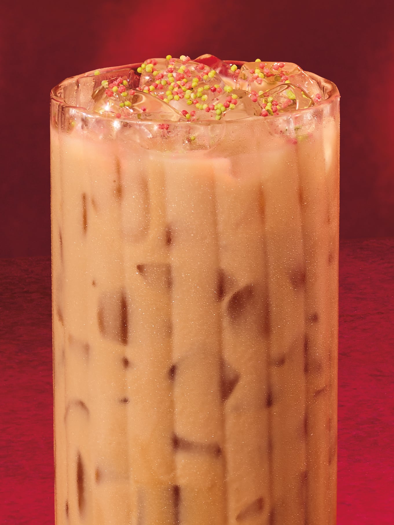 A tall glass filled with iced beverage, featuring ice cubes and colorful sprinkles on top, set against a red background.