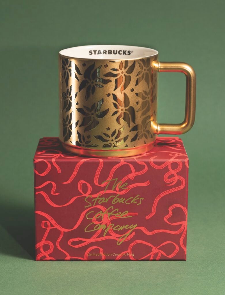 A gold ceramic mug with a floral cutout design sits atop a red box featuring playful, swirling patterns and green text.