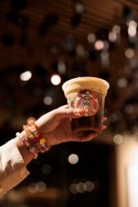 A hand holds a Starbucks cup filled with iced coffee topped with foam. Colorful beaded bracelets adorn the wrist, with a blurred background.