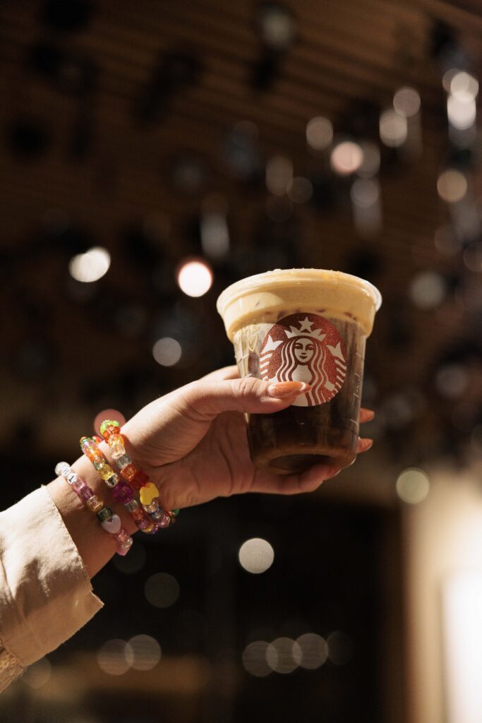 A hand holds a Starbucks cup filled with iced coffee topped with foam. Colorful beaded bracelets adorn the wrist, with a blurred background.
