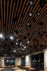 A modern café interior features a wooden ceiling with numerous hanging circular elements, some displaying a logo. Tables and chairs are arranged below, with large windows and artwork on the walls.
