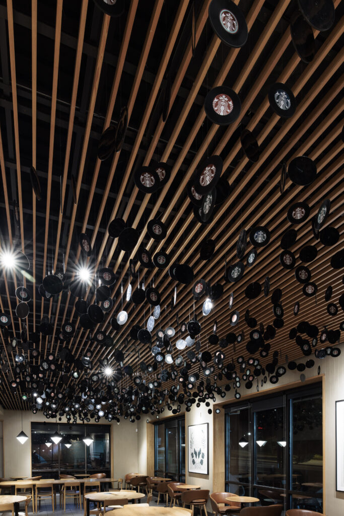 A modern café interior features a wooden ceiling with numerous hanging circular elements, some displaying a logo. Tables and chairs are arranged below, with large windows and artwork on the walls.