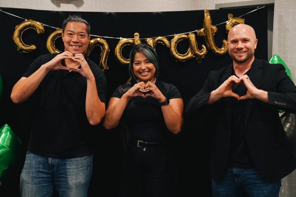Three individuals are standing together, each making a heart shape with their hands. They are smiling and dressed in black attire, with a gold "Congrats" banner in the background.