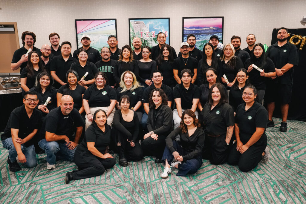 A large group of people poses together, all wearing black shirts. They are arranged in a semi-circle with a backdrop featuring colorful framed images of Texas landscapes. The setting appears to be a conference or meeting space.