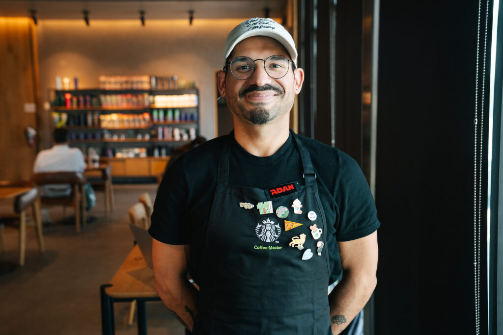 A smiling barista stands in a coffee shop, wearing a black apron adorned with various pins, including a Starbucks logo. He has glasses and a white cap. The background features shelves filled with colorful coffee products.