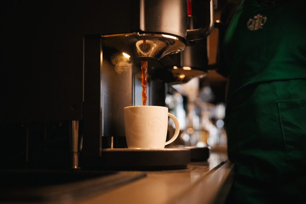 A white coffee cup with a logo is positioned under a coffee machine, as hot coffee is being poured into it. The background features a blurred café setting.