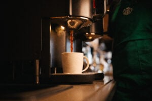 A white coffee cup with a logo is positioned under a coffee machine, as hot coffee is being poured into it. The background features a blurred café setting.