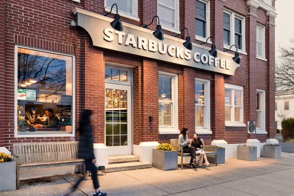 A Starbucks coffee shop with a brick exterior features two women sitting on a bench outside, enjoying drinks. A person walks by.
