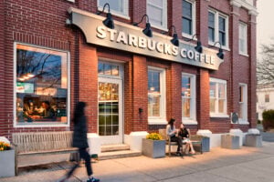 A Starbucks coffee shop with a brick exterior features two women sitting on a bench outside, enjoying drinks. A person walks by.