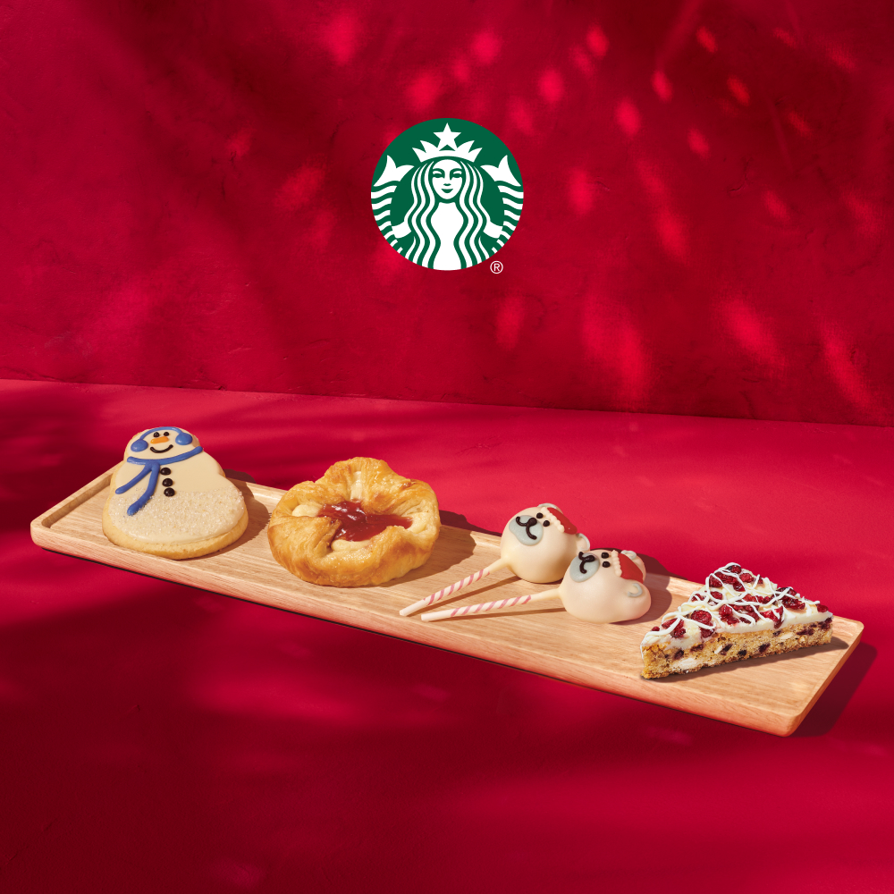 A wooden tray holds festive treats: a snowman cookie, a fruit-filled pastry, two decorated cake pops, and a slice of dessert topped with red icing. The background is a vibrant red.