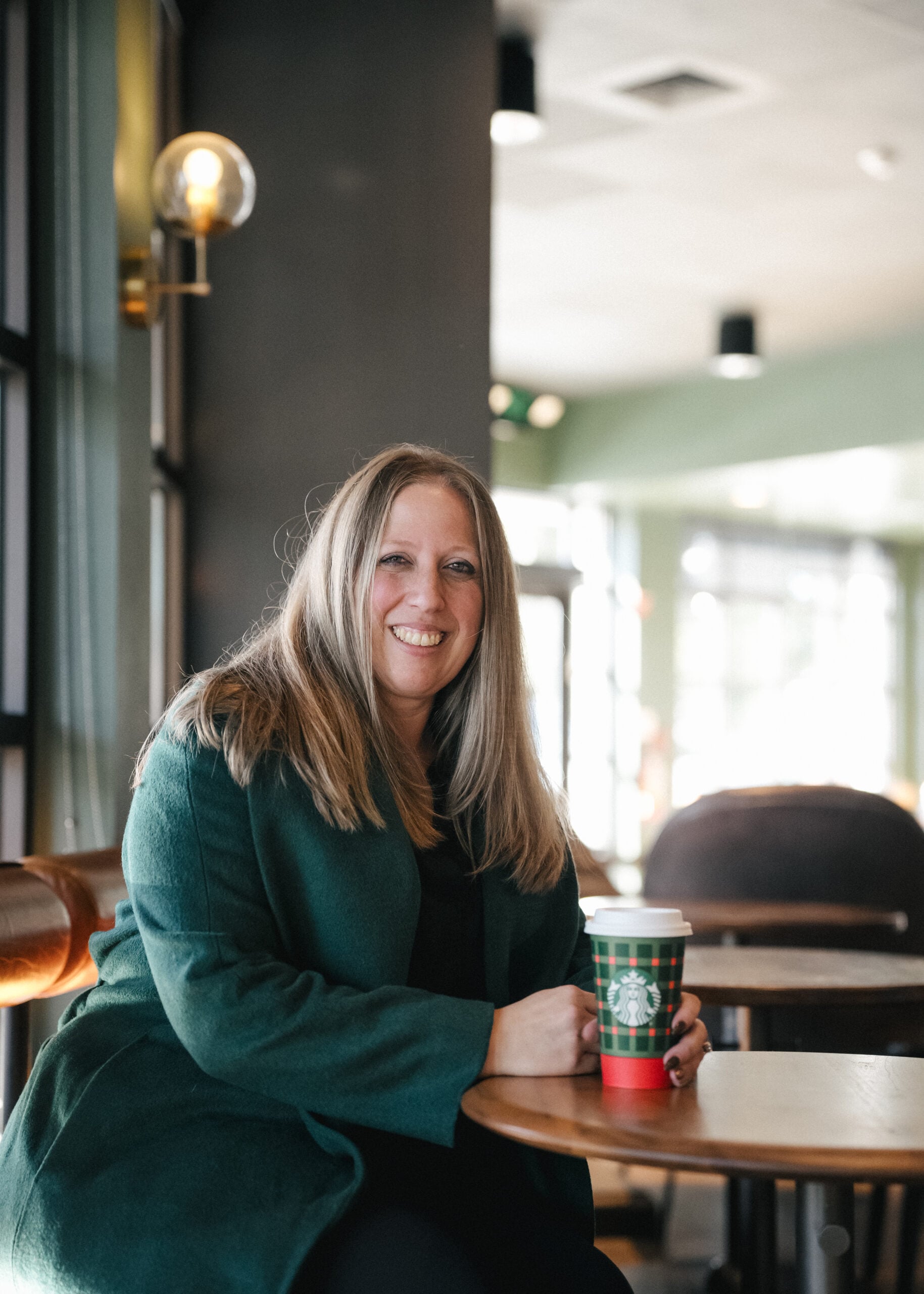A woman with long, straight hair sits at a table in a café, wearing a green coat. She holds a festive Starbucks cup and smiles.