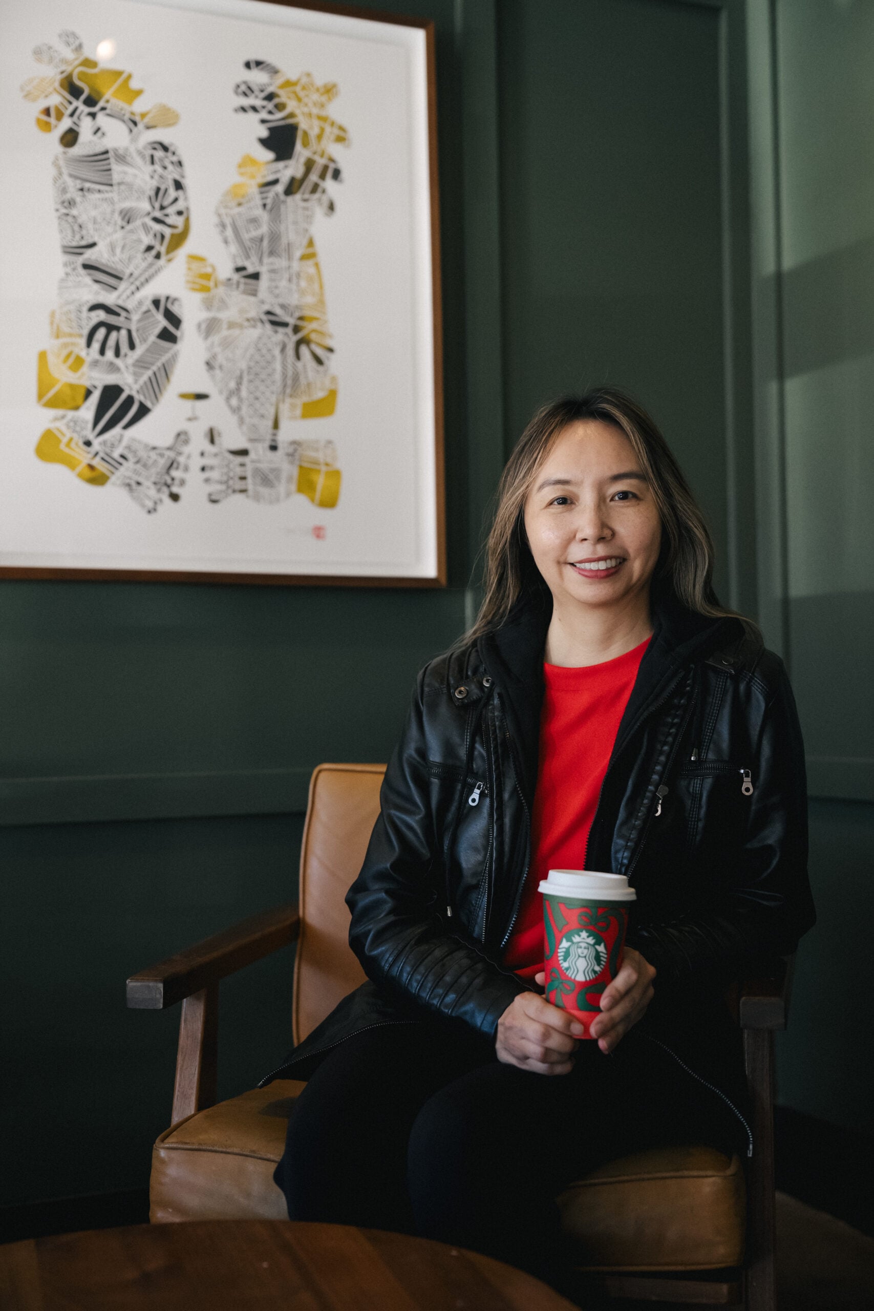 A woman sits in a cozy café, wearing a black leather jacket over a red shirt, holding a Starbucks cup. Behind her is a colorful artwork on the green wall.