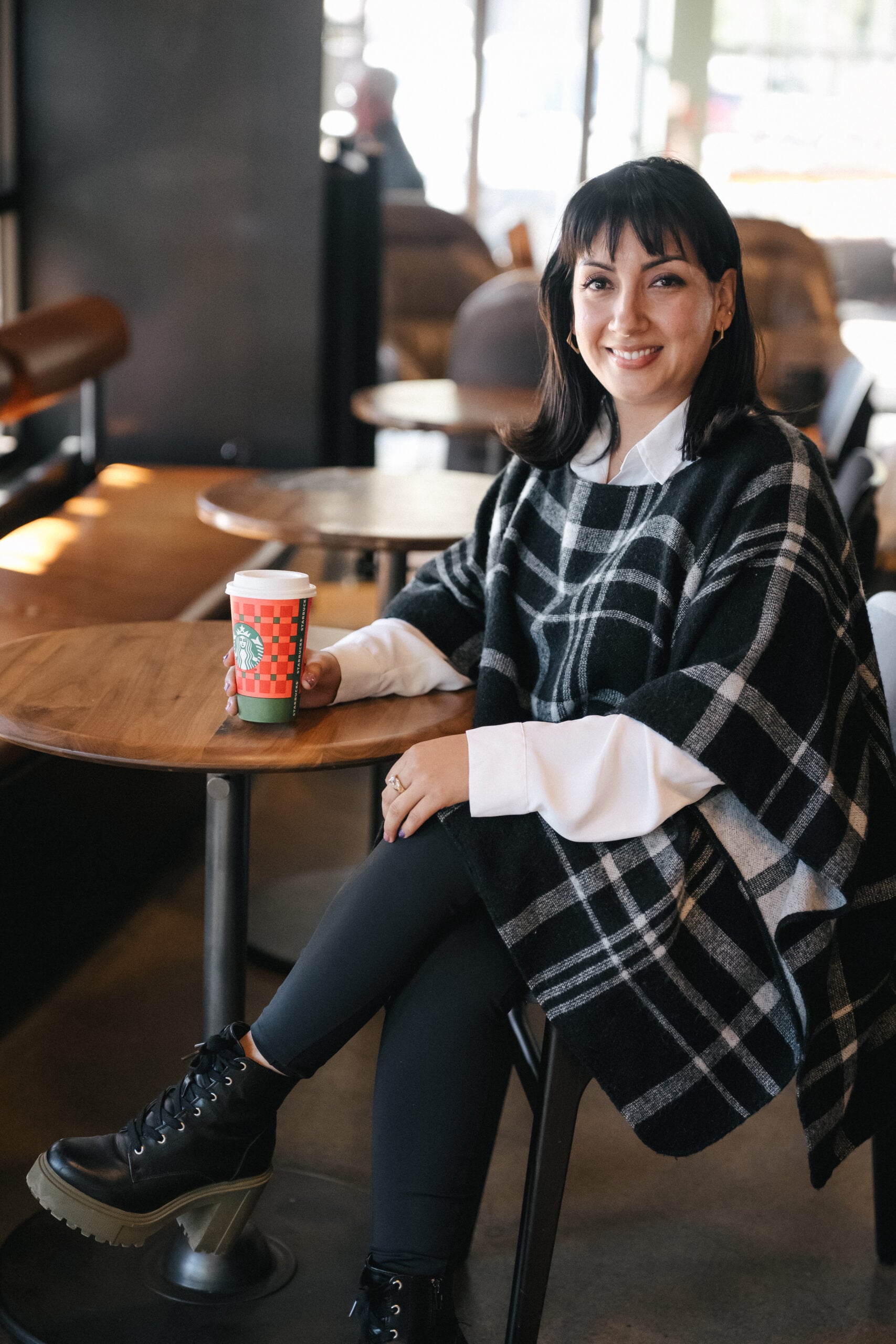 A woman sits at a café table, wearing a black and white plaid poncho over a white shirt. She holds a Starbucks cup and smiles.