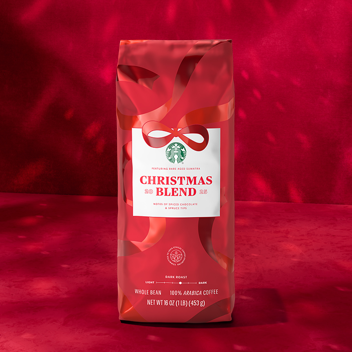A festive red coffee bag labeled "Christmas Blend" features a white panel with text detailing flavor notes of spiced chocolate and spruce tips. The bag is designed with a ribbon motif and includes the Starbucks logo.