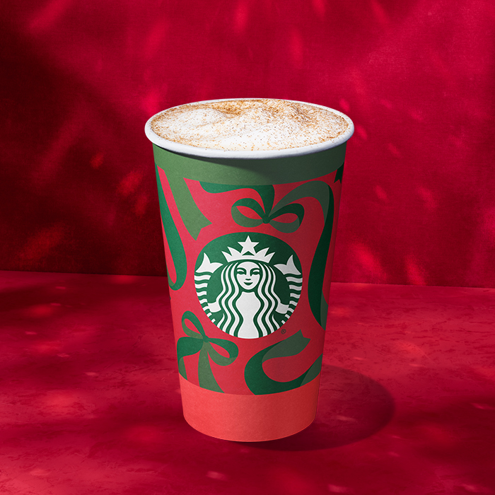 A festive Starbucks cup featuring a green and red design with holiday motifs, topped with frothy milk and cinnamon.