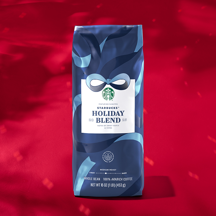 A blue and white coffee bag labeled "Starbucks Holiday Blend," featuring notes of white maple and herbs. It contains whole bean, medium roast coffee. The background is a vibrant red.