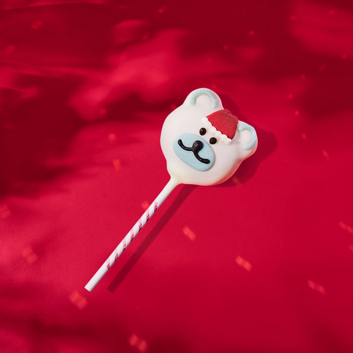 A cute polar bear-shaped lollipop with a red Santa hat, featuring a blue nose and black eyes, set against a red background.