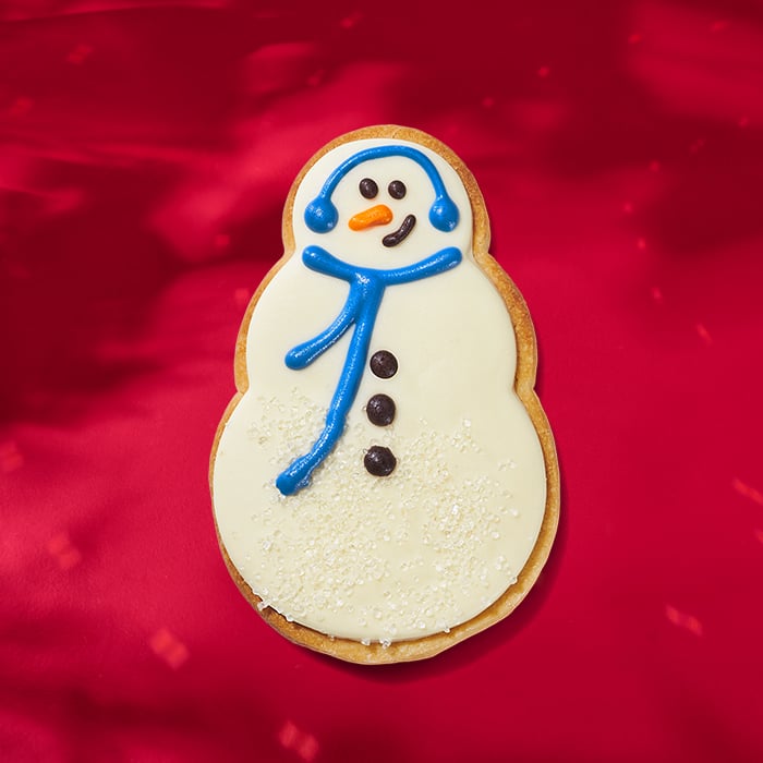 A decorated cookie shaped like a snowman, featuring white icing, blue scarf and earmuffs, and orange nose, on a red background.
