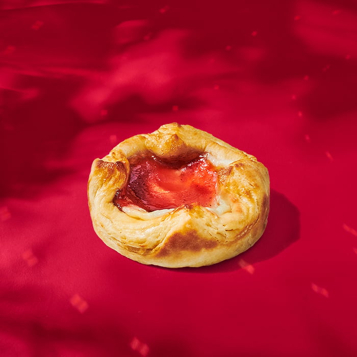 A golden-brown pastry with a flaky texture, featuring a glossy red fruit filling at the center, set against a vibrant red background.