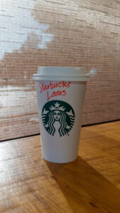 A Starbucks coffee cup with a green logo sits on a wooden table. The cup has "Starbucks Lovers" written in red on the side.