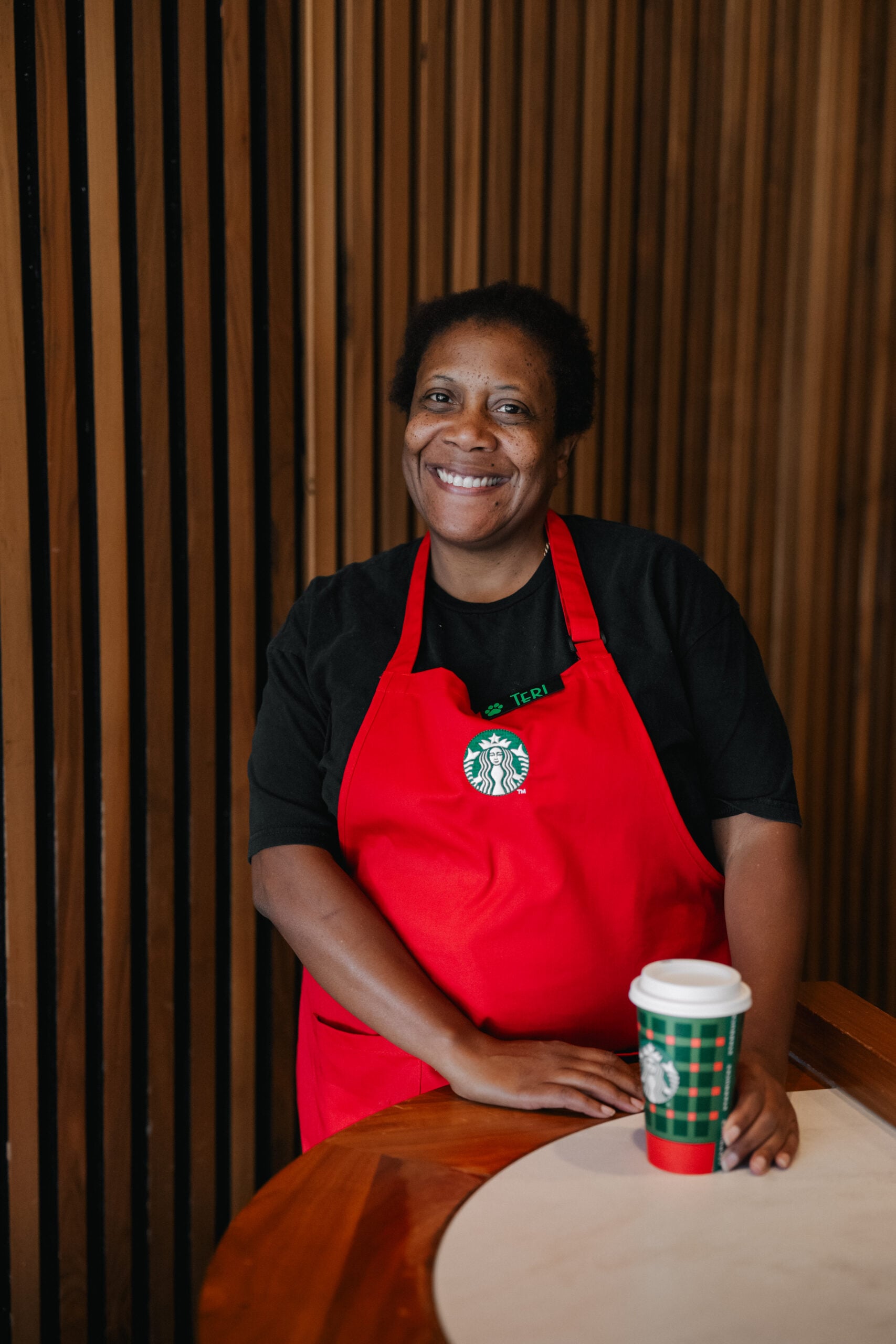 A smiling barista in a red apron stands by a wooden table, holding a festive Starbucks cup. The background features wooden slats.