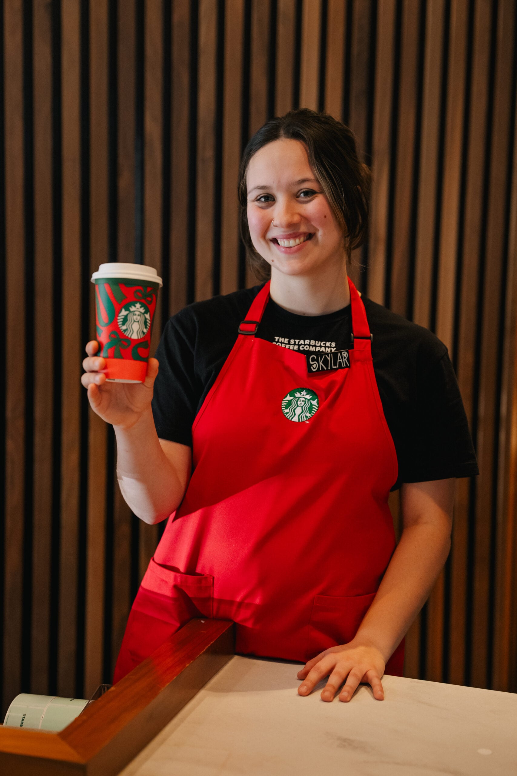 A barista in a red apron smiles while holding a decorated coffee cup. The background features wooden paneling and a counter.