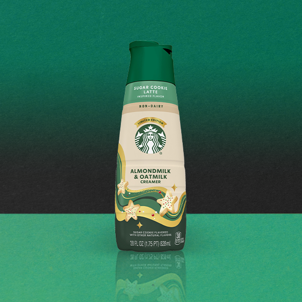A bottle of Starbucks Almondmilk & Oatmilk Creamer, featuring a sugar cookie latte flavor. The design includes festive star motifs and a green cap.