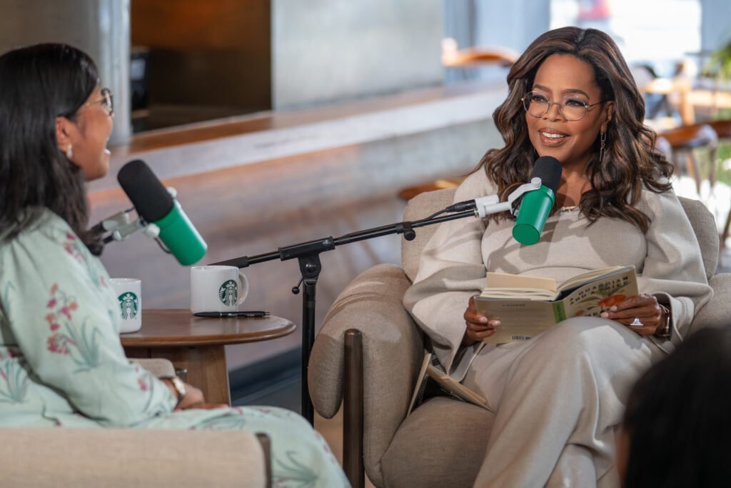 A woman with long, wavy hair sits in a cozy chair, holding a book and smiling. She wears a light-colored outfit and glasses. Two microphones are positioned in front of her, and there are Starbucks mugs on a small table nearby. The setting appears to be a relaxed, modern café.