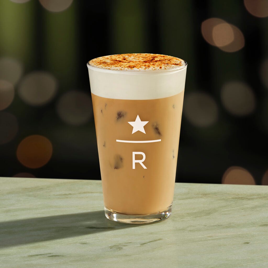 A clear glass filled with iced coffee topped with frothy cream and a sprinkle of cinnamon, featuring a logo with a star and "R.