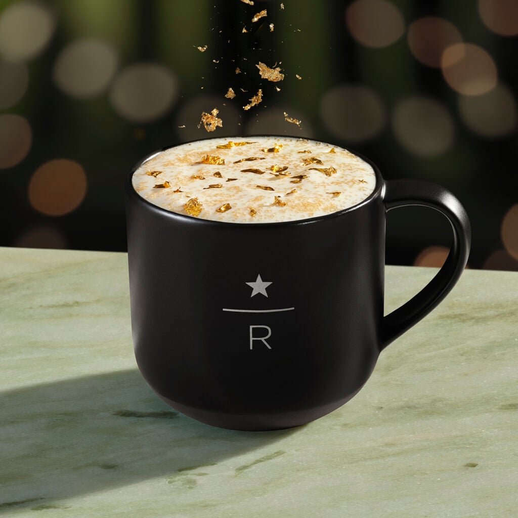 A black mug with a star and "R" design holds a creamy beverage topped with golden flakes, set on a marble surface.