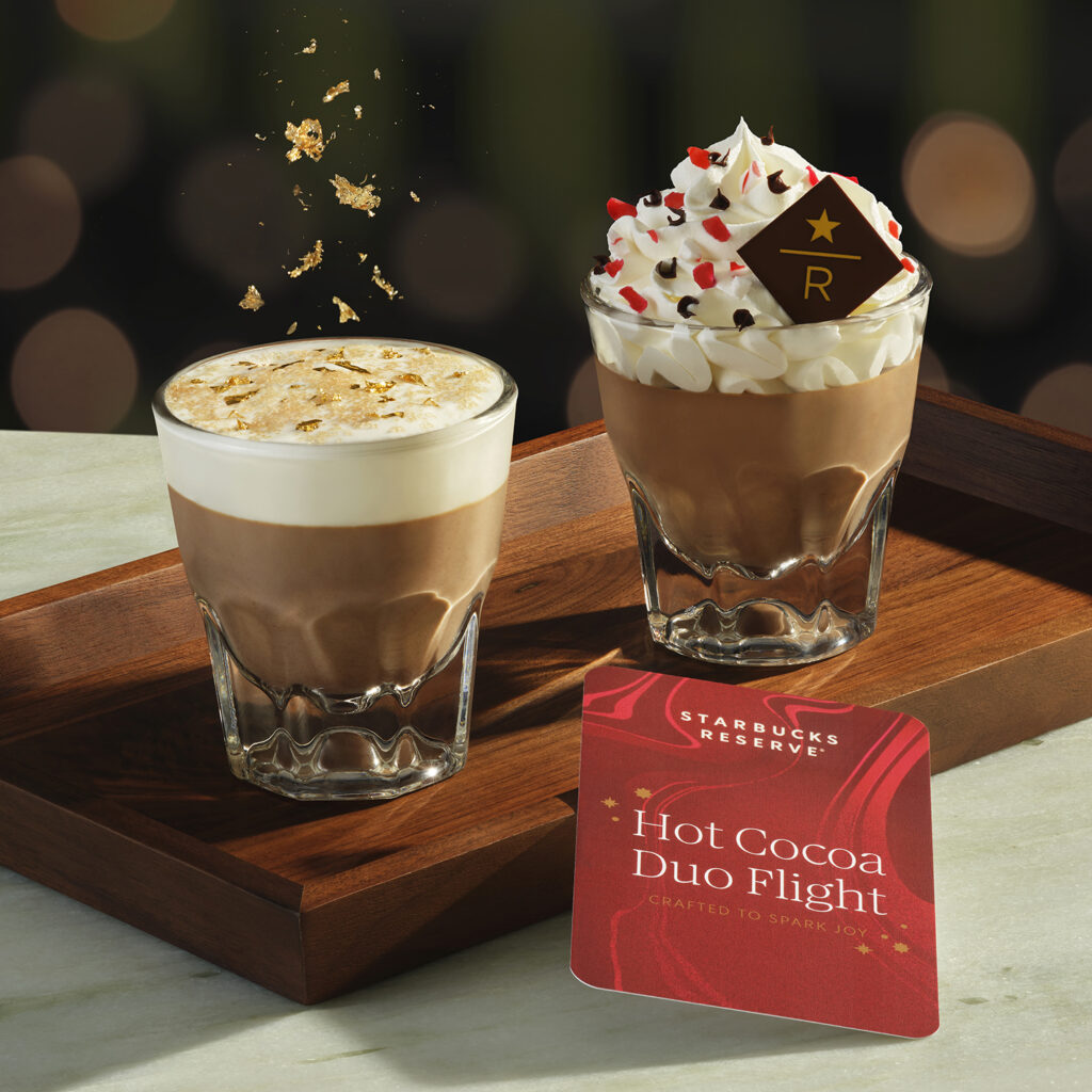 Two glasses of hot cocoa are presented on a wooden tray. One has a creamy top with gold flakes, while the other is topped with whipped cream, chocolate, and red sprinkles, featuring a chocolate emblem. A card labeled "Hot Cocoa Duo Flight" is placed beside them.
