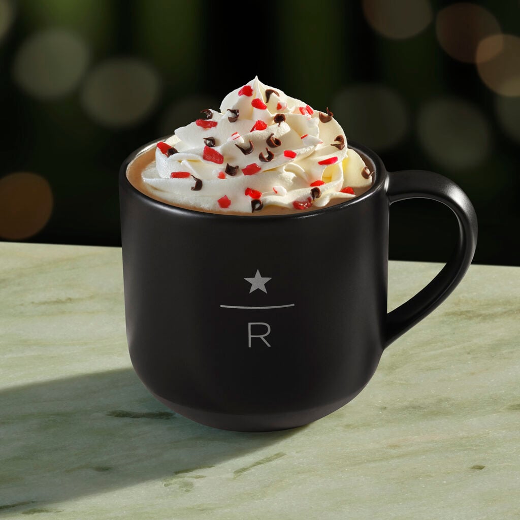 A black mug with a star and "R" design holds a creamy beverage topped with whipped cream, red sprinkles, and chocolate shavings.