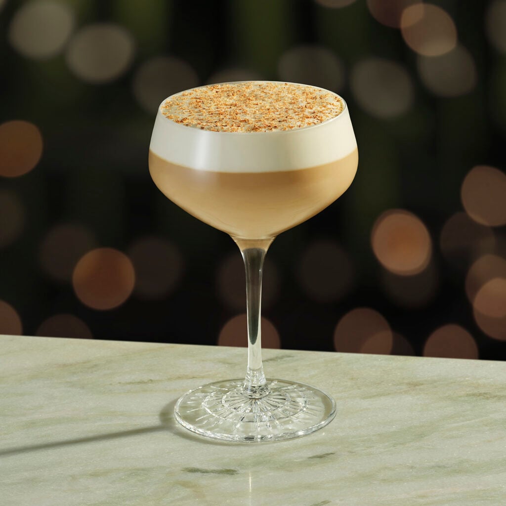 A stylish cocktail in a coupe glass, featuring a layered design with a creamy top and a dusting of spices, set on a marble surface.