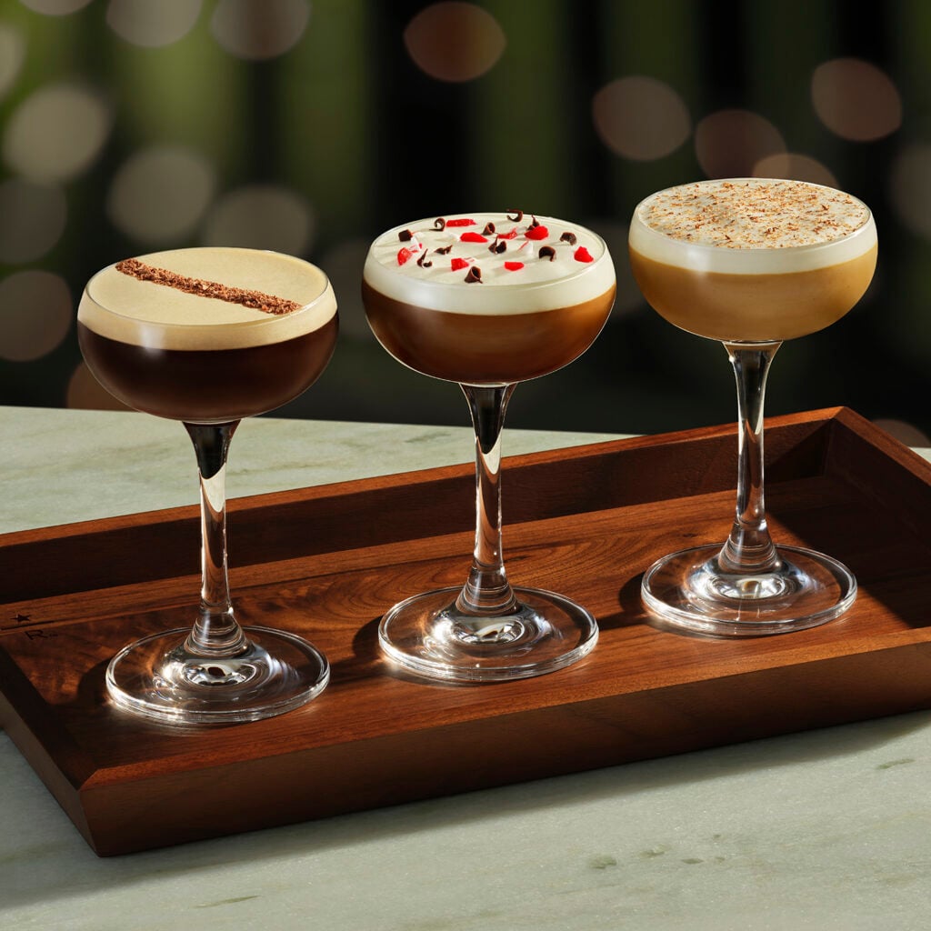 Three elegant cocktails are displayed on a wooden tray. The left cocktail features a dark layer topped with a light foam and a chocolate garnish. The middle drink has a dark base with a creamy layer adorned with red and white sprinkles. The right cocktail showcases a light brown base with a frothy top and a sprinkle of nutmeg or cinnamon. The background is softly blurred, enhancing the focus on the drinks.