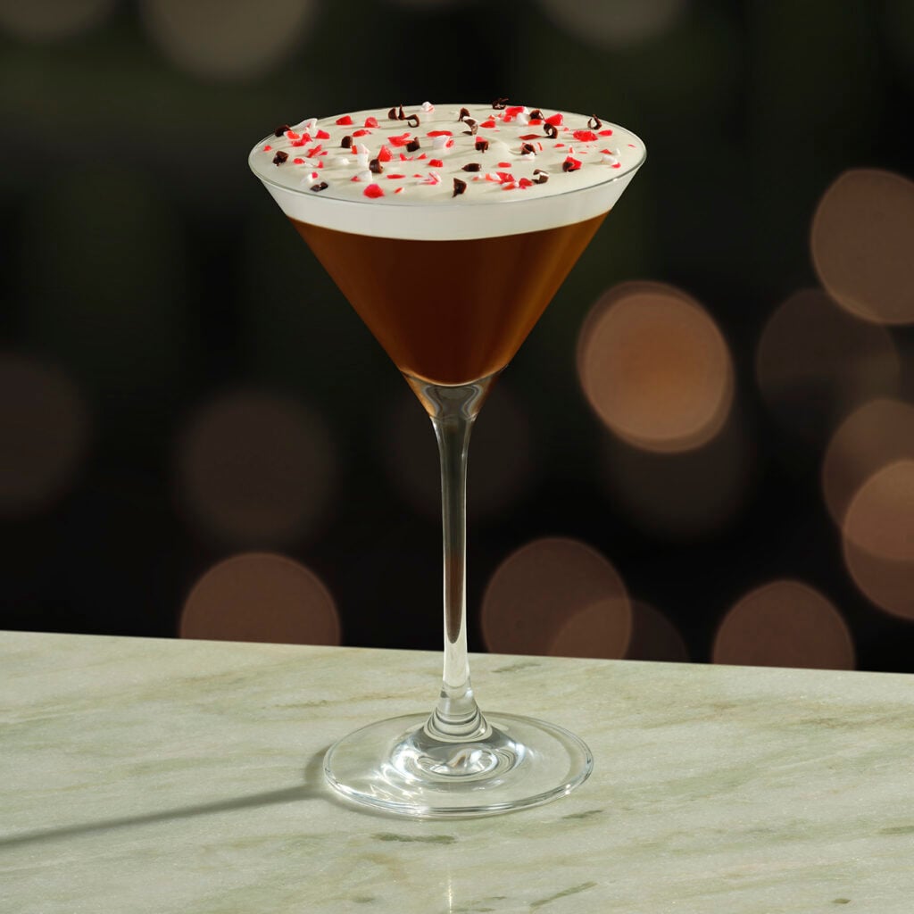 A cocktail in a tall, elegant glass features a dark liquid topped with whipped cream, red and white sprinkles, and chocolate shavings. The background is softly blurred with warm bokeh lights.