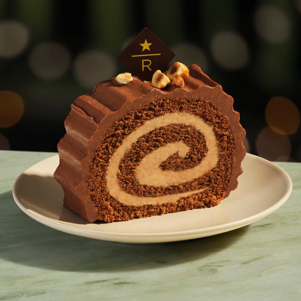 A slice of chocolate roll cake with a creamy filling, topped with a chocolate decoration and crushed nuts, served on a plate.