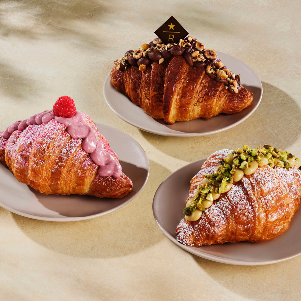 Three decorative croissants are displayed on beige plates. The left croissant is topped with pink frosting and a raspberry. The center croissant features chocolate frosting and chopped hazelnuts. The right croissant is adorned with pistachios and a dusting of powdered sugar.