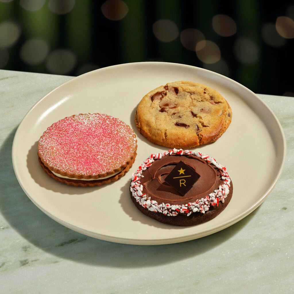 A beige plate holds three cookies: a pink-sprinkled sandwich cookie, a chocolate chip cookie, and a chocolate frosted cookie with peppermint bits.