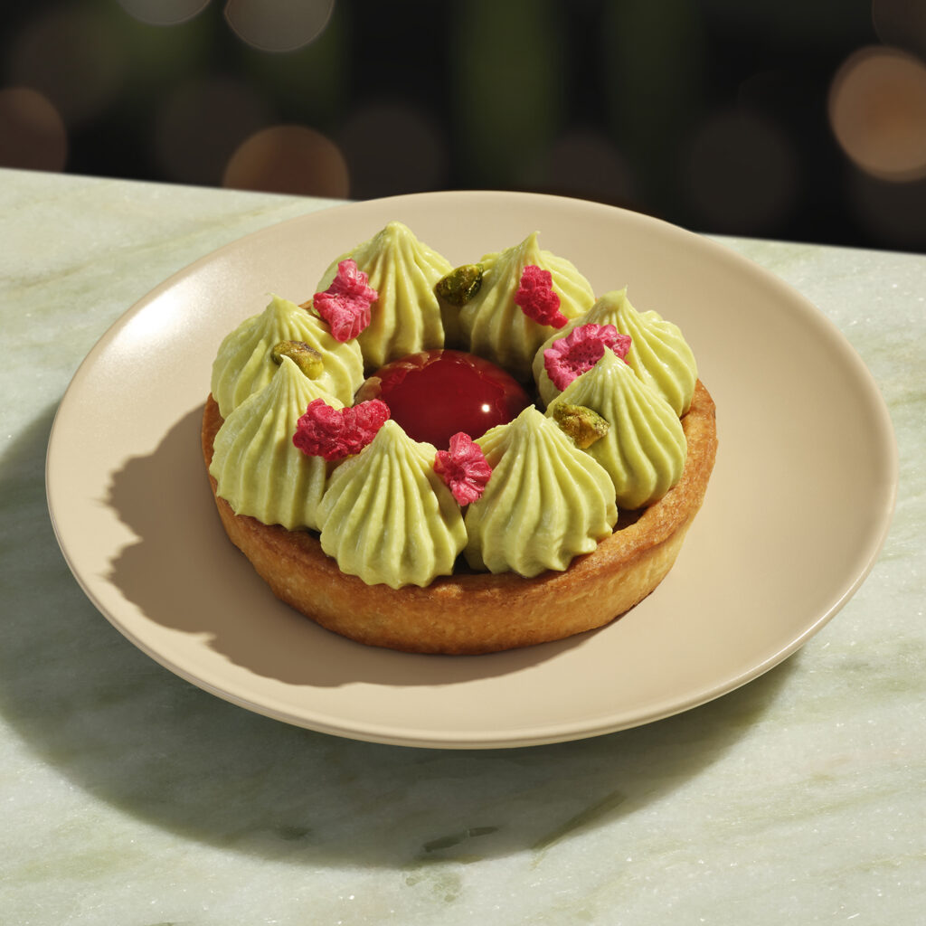 A round pastry tart topped with green piped frosting, a glossy red center, and scattered pink and green decorations on a beige plate.