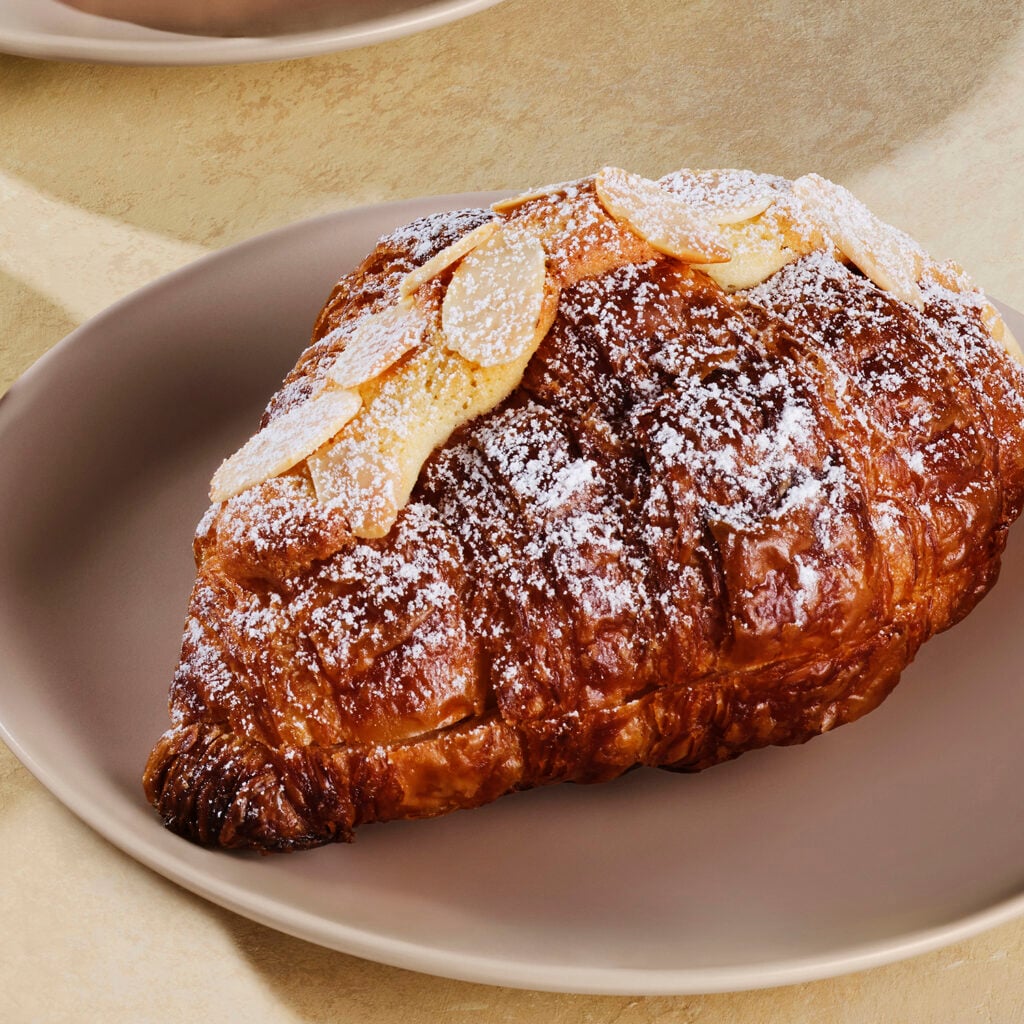 A golden-brown almond croissant topped with sliced almonds and a dusting of powdered sugar, served on a light-colored plate.