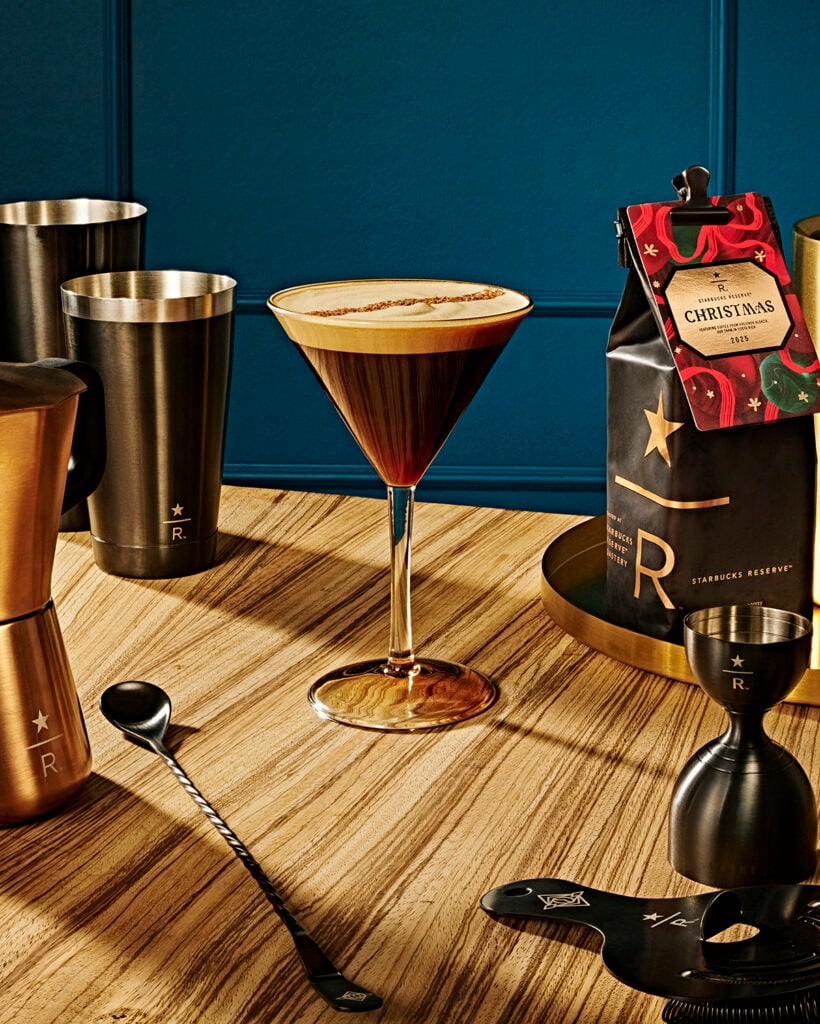 A stylish cocktail glass filled with a dark beverage topped with foam sits on a wooden table. Nearby are various coffee tools, including a measuring jigger, a spoon, and a bag of Starbucks Reserve Christmas coffee. The background features a deep blue wall, enhancing the warm tones of the wooden surface and metallic drinkware.