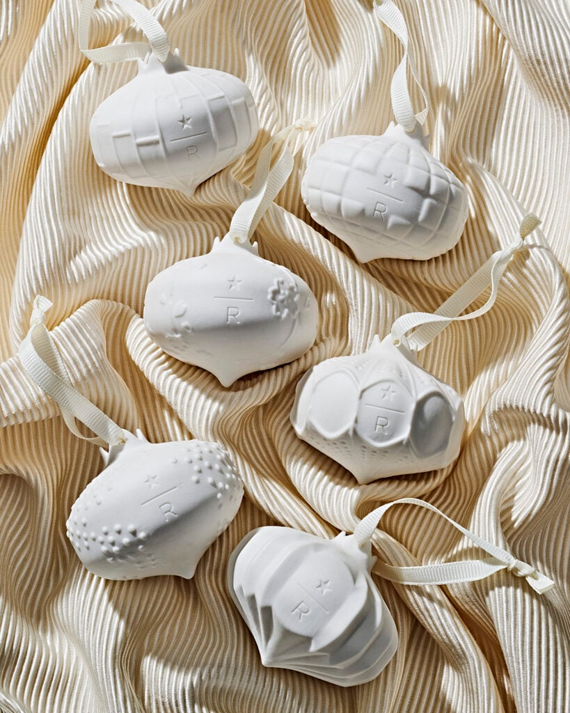 Six white ceramic ornaments with various textures and shapes are arranged on a soft, cream-colored fabric. Each ornament features a ribbon for hanging and has a small star and letter "R" embossed on them.