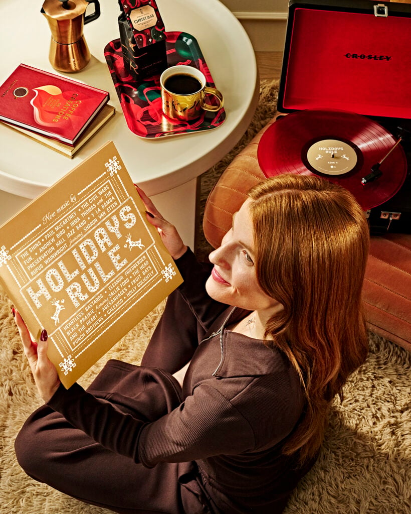 A woman with long red hair sits on a plush rug, holding a gold album cover titled "HOLIDAY'S RULE." Nearby, a coffee cup, a festive tray, and a record player are visible.