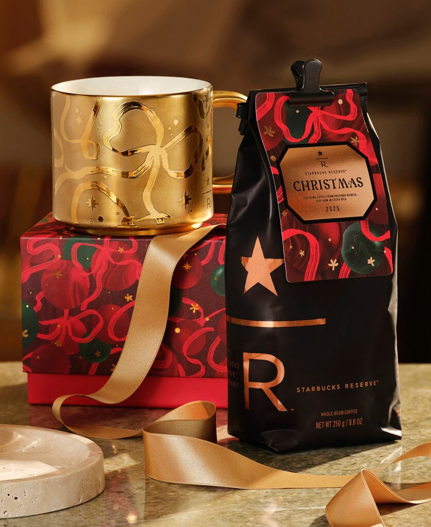 A gold mug with a decorative design sits next to a black bag of Starbucks Reserve Christmas coffee, placed on a festive gift box with a gold ribbon.