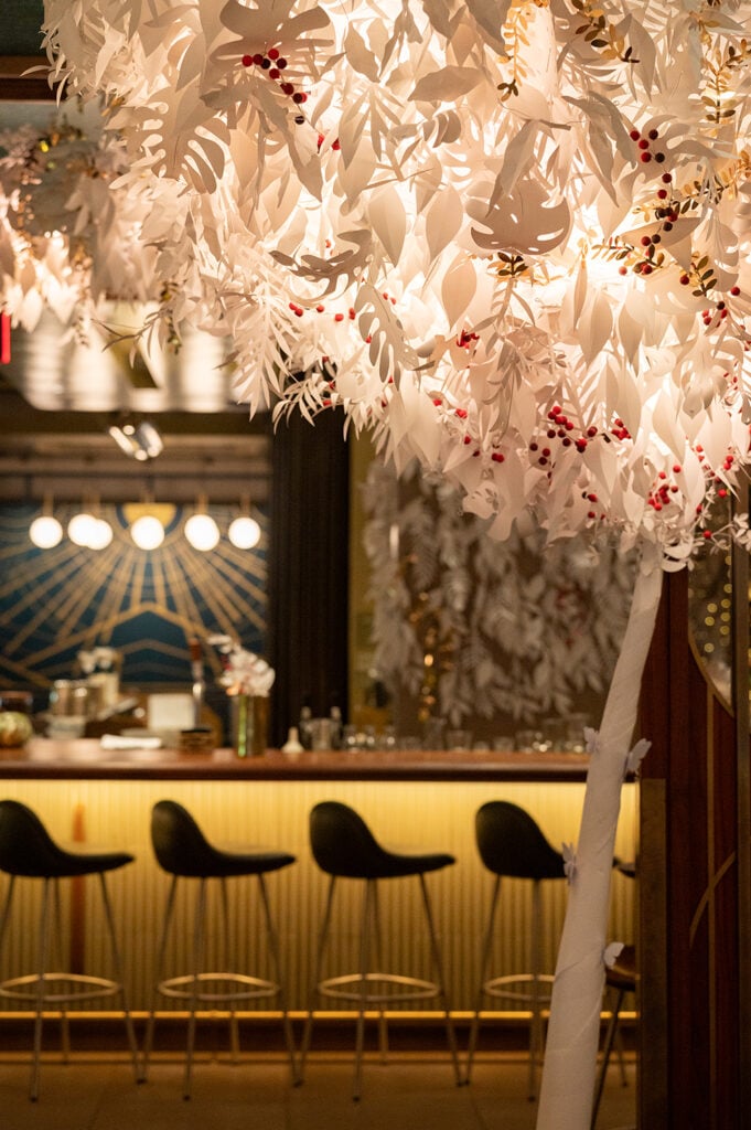 A beautifully decorated restaurant interior featuring a large, intricate light fixture made of white paper leaves and red berries. The bar area is visible in the background, with stylish black stools and warm lighting.