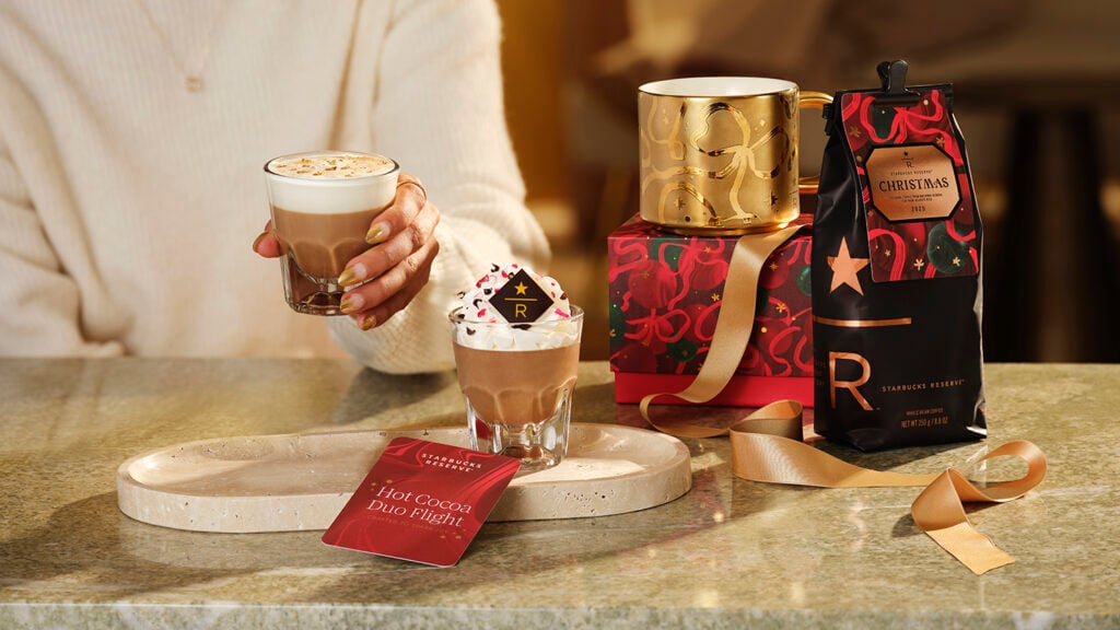 A hand holds a glass of hot cocoa topped with whipped cream and sprinkles. Nearby, a gold mug and a festive coffee bag are displayed, along with a card reading "Hot Cocoa Duo Flight." The setting features a marble surface and warm lighting.