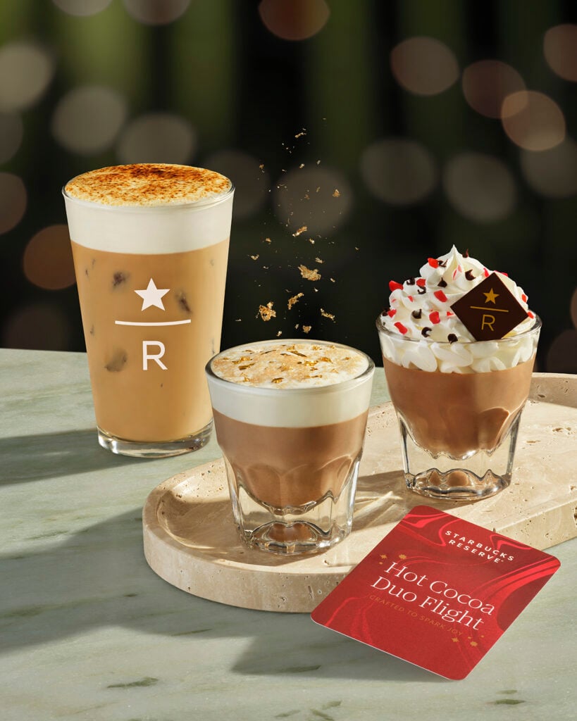 Three coffee beverages are displayed on a light stone tray. On the left, an iced coffee with a frothy top. In the center, a hot cocoa topped with whipped cream and festive sprinkles. To the right, a smaller glass of hot cocoa with a caramelized top. A red card labeled "Hot Cocoa Duo Flight" is placed beside the drinks. The background features soft, blurred lights.