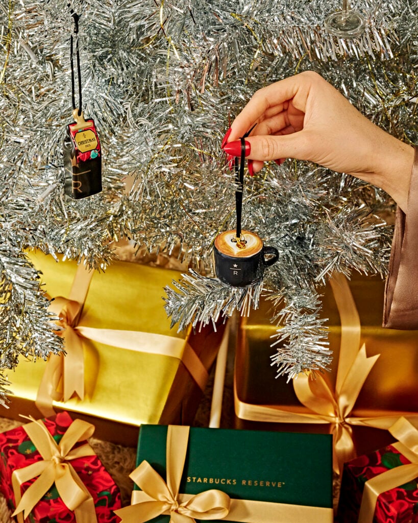 A hand with red nails hangs a miniature coffee cup ornament on a silver tinsel Christmas tree. Nearby, colorful wrapped gifts are visible.